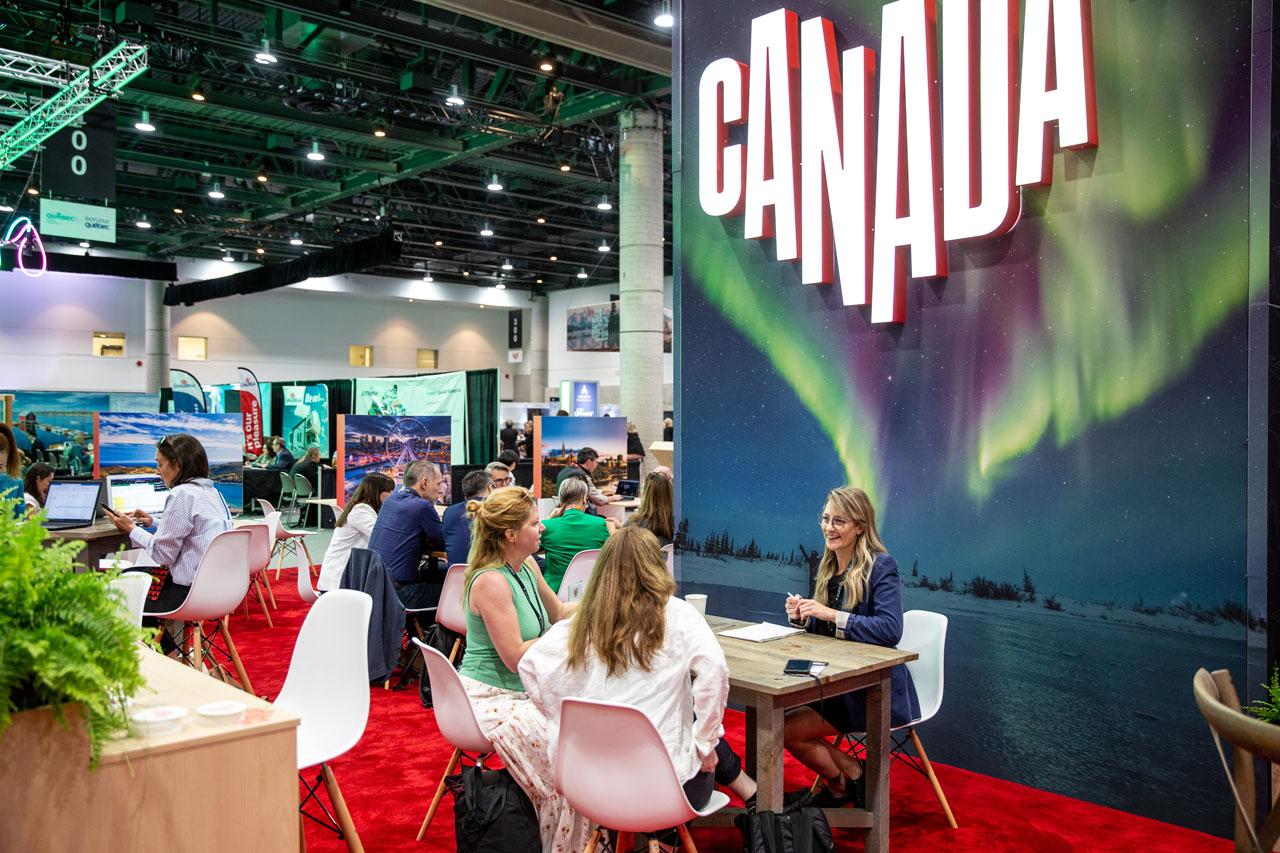 Des gens sont assis à des tables. Derrière eux, il y a un grand logo Canada superposé sur une image d’aurore boréale.